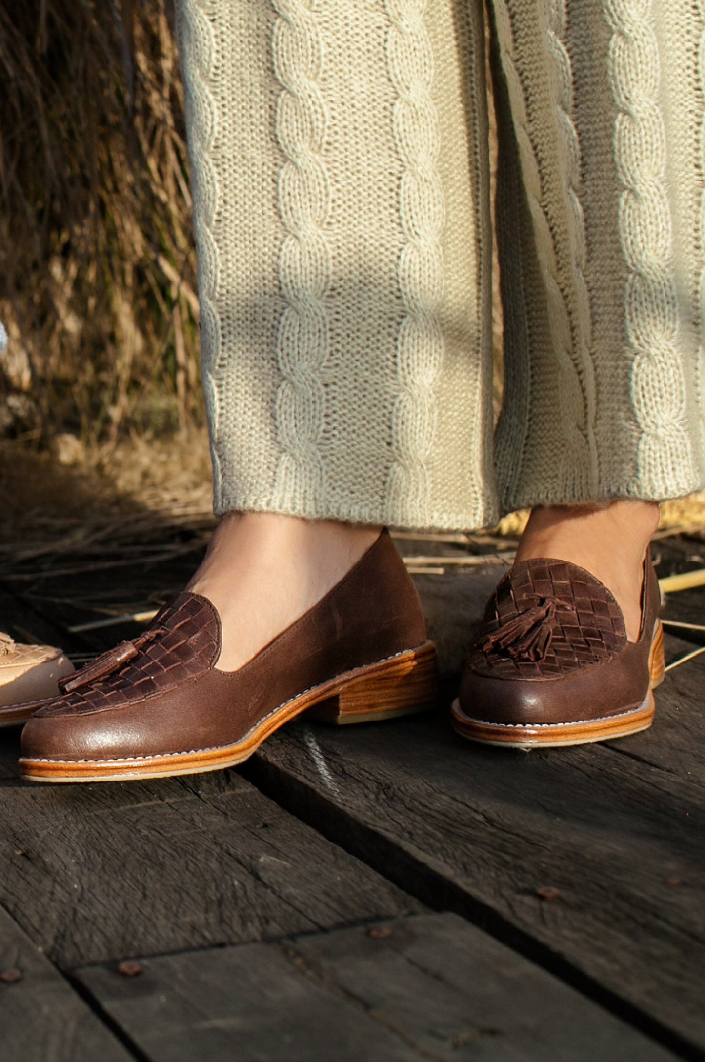 Brown loafers worn with light green knitted pants on a wooden floor.