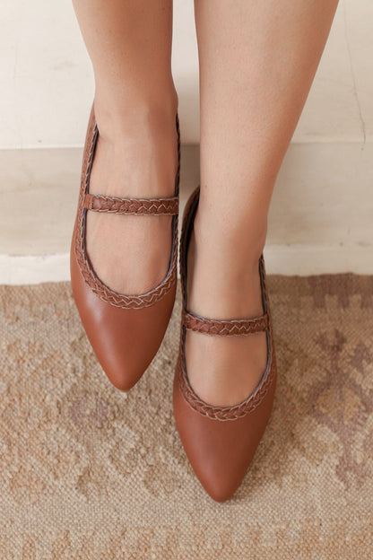 Brown pointed-toe shoes with braided details worn by a person on a textured floor.