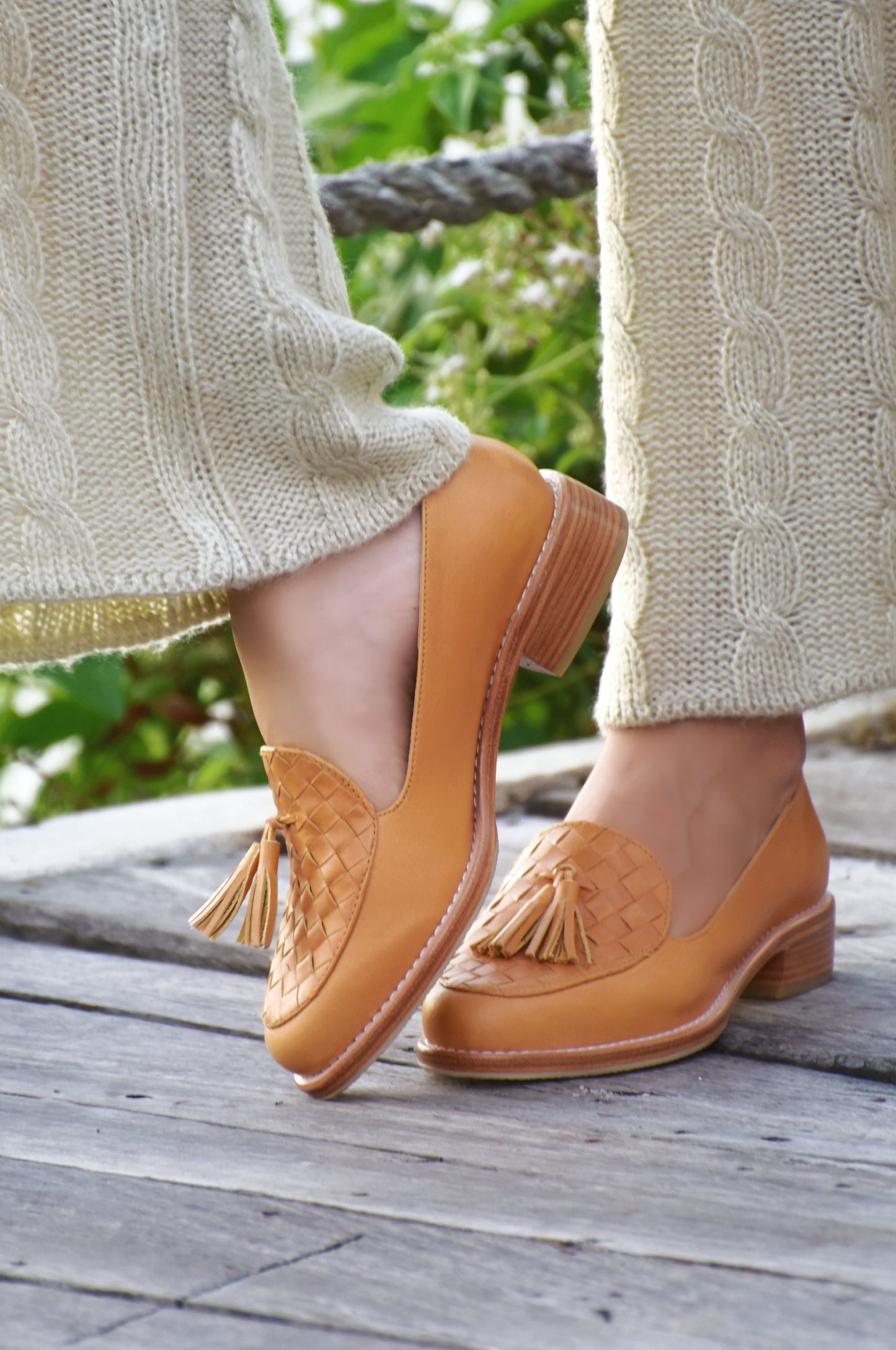 Tan loafers with tassels worn by a person on a wooden deck with greenery in the background