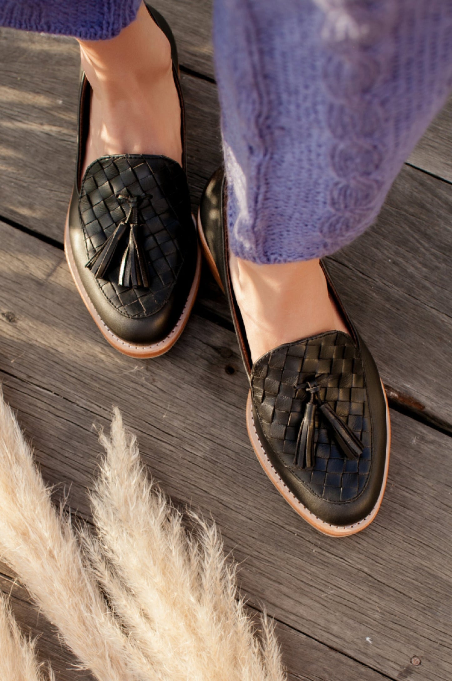 Woven loafers with tassels worn by a person on a wooden floor.