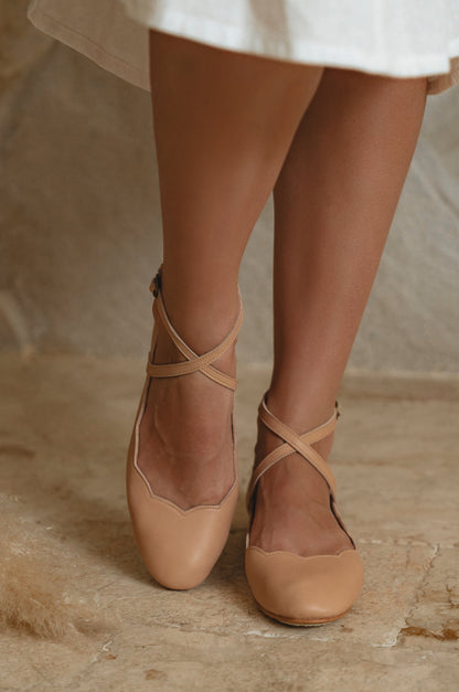 Tan ballet flats worn with a white skirt on a neutral background