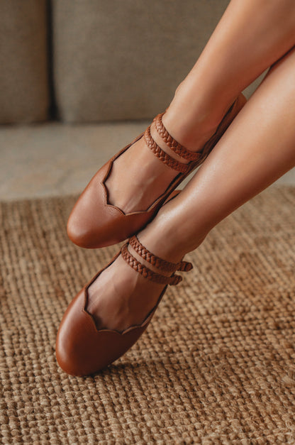 Brown leather shoes with braided straps worn by a person on a textured surface.